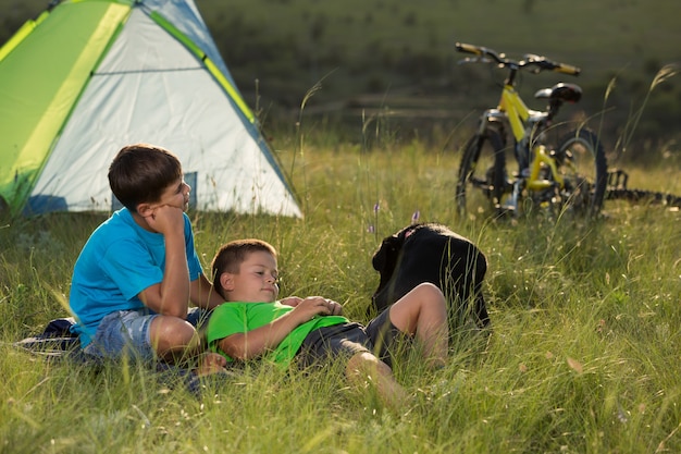 deux garçons se reposant allongés sur l'herbe avec une tente et des vélos en arrière-plan