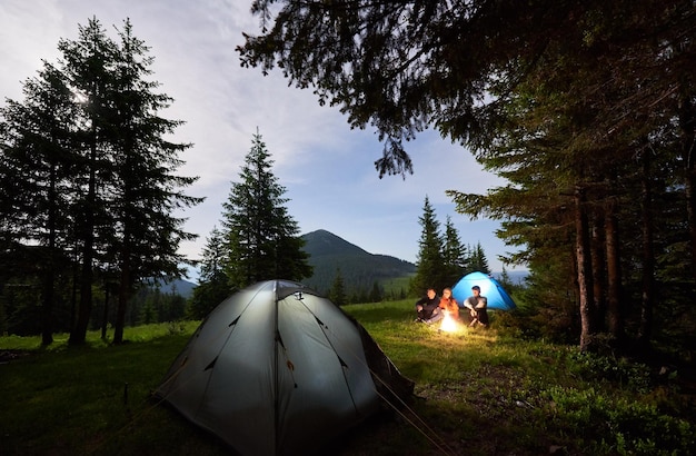 Deux gars et une fille se prélassent près d'un feu de camp dans un camping autour de pins contre des montagnes sous le ciel du soir