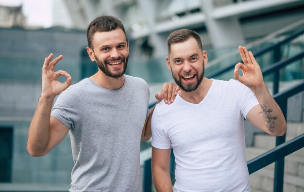 Deux heureux meilleurs amis barbus modernes excités dans des vêtements décontractés s'amusant et montrant le signe ok sur la caméra à l'extérieur.