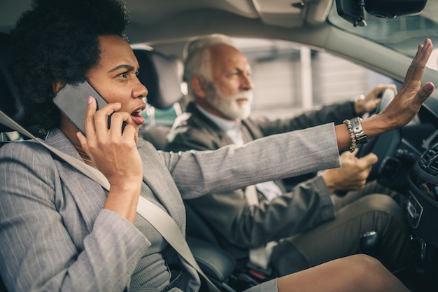 Deux hommes d'affaires multiethniques prospères voyageant en voiture. Homme senior conduisant une voiture, femme noire parlant sur un smartphone et effrayée en regardant à travers le pare-brise.