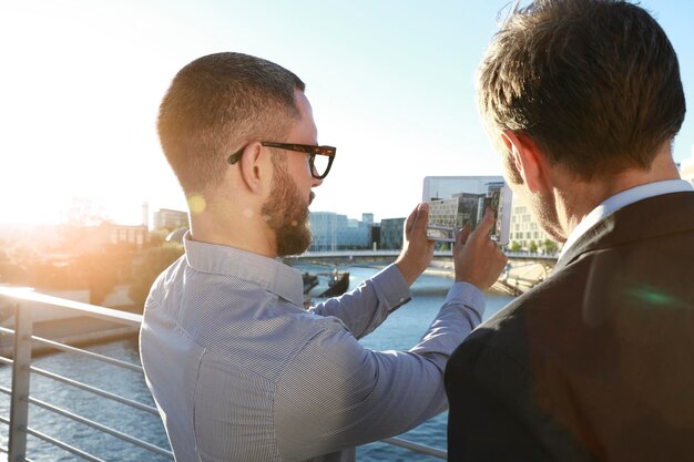 Deux hommes d'affaires prenant une photo avec une tablette sur un pont de la ville