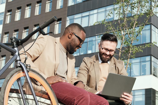 Deux hommes assis à l'extérieur dans la ville en regardant quelque chose sur un ordinateur portable et en riant