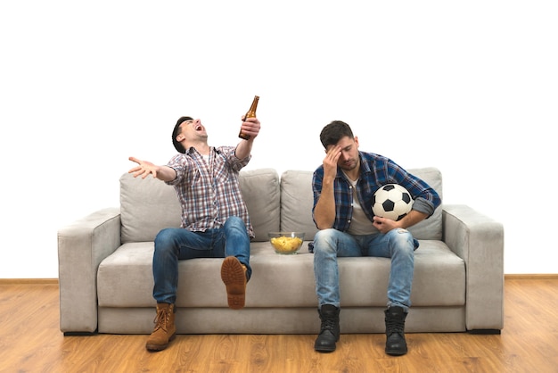 Les deux hommes avec une bière et des frites regardent un football sur le canapé