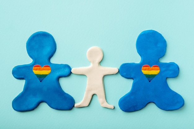 Deux hommes identiques en pâte à modeler bleue avec des formes de coeur arc-en-ciel en tant que parents de même sexe tenant la main d'un enfant en pâte à modeler blanche sur fond bleu. Notion de famille LGBT. Notion de FAMILLES DIVERSIFIÉES.