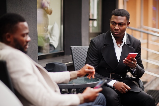 Deux hommes noirs de mode assis en plein air avec un téléphone mobile. Portrait à la mode des modèles masculins afro-américains. Portez un costume, un manteau et un chapeau.