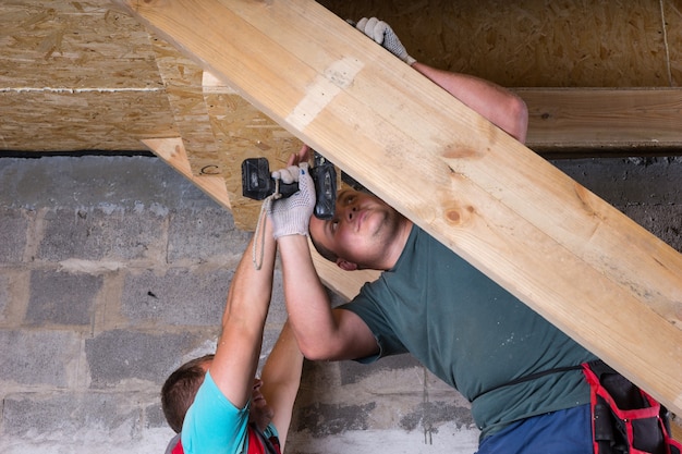 Deux hommes travaillant ensemble pour construire un cadre en bois pour les escaliers avec des outils électriques, menant au sous-sol d'une maison inachevée