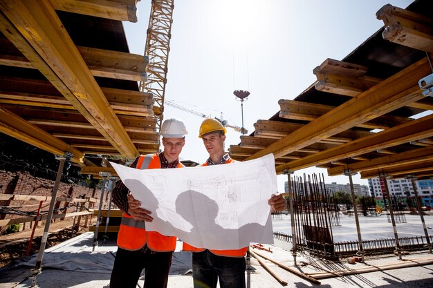 Deux hommes vêtus de chemises, de gilets de travail orange et de casques explorent la documentation de construction sur le chantier près des constructions en bois .