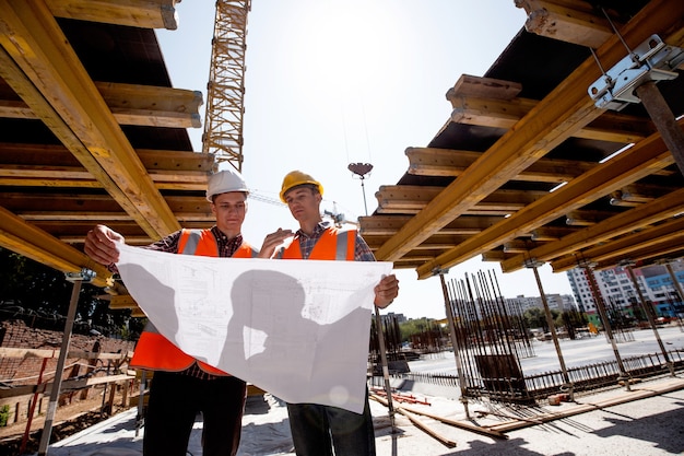 Deux hommes vêtus de chemises, de gilets de travail orange et de casques explorent la documentation de construction sur le chantier près des constructions en bois .
