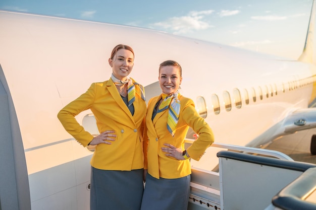 Deux hôtesses de l'air joyeuses en uniforme de compagnie aérienne regardant la caméra et souriant tout en se tenant près de la porte de l'avion