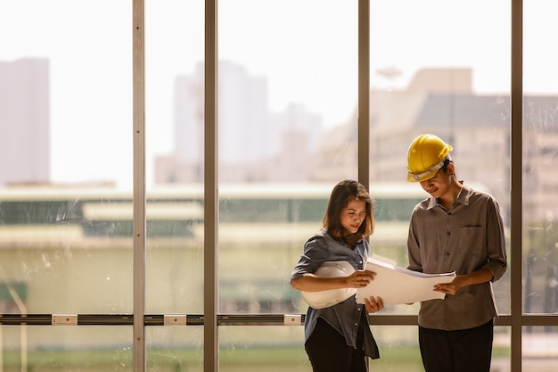 Deux ingénieurs asiatiques, un homme avec un casque de sécurité jaune et une femme avec un blanc debout et parlant près d'un haut cadre en verre de mur-rideau sur un chantier de construction. Les deux regardent le papier de plan.