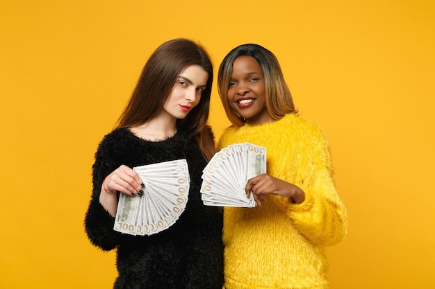 Deux jeunes amies amusantes européennes et africaines en vêtements jaunes noirs tenant un ventilateur d'argent liquide en billets en dollars isolés sur fond de mur orange. Concept de style de vie des gens. Maquette de l'espace de copie.
