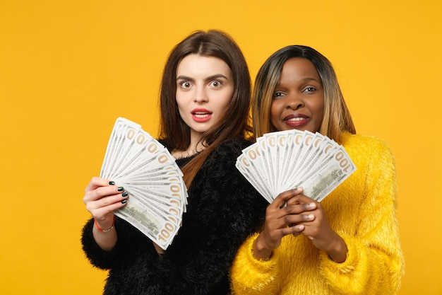 Deux jeunes amies amusantes européennes et africaines en vêtements jaunes noirs tenant un ventilateur d'argent liquide en billets en dollars isolés sur fond de mur orange. Concept de style de vie des gens. Maquette de l'espace de copie.