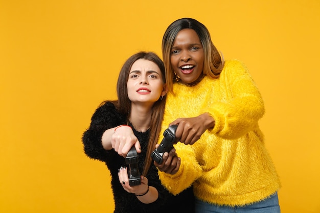 Deux jeunes amies amusantes européennes et afro-américaines en vêtements jaunes noirs tiennent le joystick isolé sur fond de mur orange vif, portrait en studio. Concept de style de vie des gens. Maquette d'espace de copie