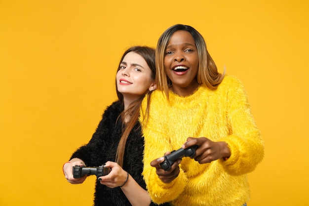 Deux jeunes amies amusantes européennes et afro-américaines en vêtements jaunes noirs tiennent le joystick isolé sur fond de mur orange vif, portrait en studio. Concept de style de vie des gens. Maquette d'espace de copie