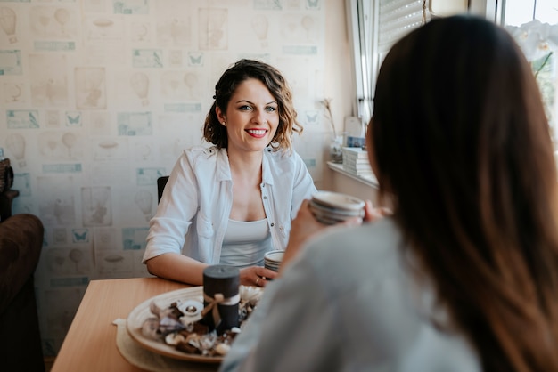 Deux jeunes amies heureux avec des tasses à café converser dans le salon à la maison.