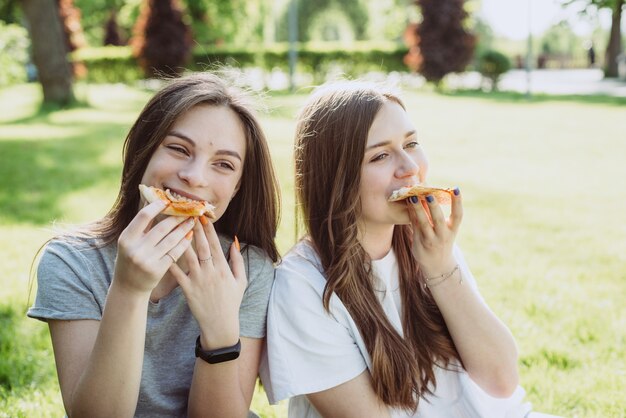 Deux jeunes amis adolescents joyeux dans le parc en train de manger de la pizza. Les femmes mangent de la restauration rapide. Pas une alimentation saine. Mise au point sélective douce.