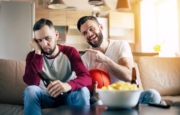 Deux jeunes amis drôles excités fans de basket-ball en regardant un match de télévision et en criant tout en se reposant sur le canapé