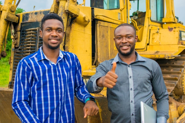 Deux jeunes beaux entrepreneurs africains se sentent dépassés alors qu'ils se tiennent à côté d'un tracteur et ont levé le pouce