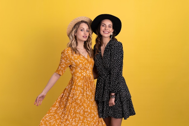 deux jeunes belles femmes amis ensemble isolés sur jaune en robe noire et jaune et chapeau boho élégant s'amusant