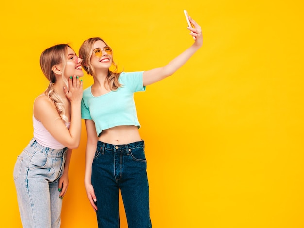 Deux jeunes belles femmes hipster blondes souriantes dans des vêtements d'été à la mode Sexy femmes insouciantes posant près du mur jaune en studio Modèles positifs s'amusant Gai et heureux Prendre des photos de selfie