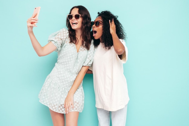 Deux jeunes belles femmes hipster internationales souriantes dans des vêtements d'été à la mode Sexy femmes insouciantes posant près du mur bleu en studio Modèles positifs s'amusant dans des lunettes de soleil Prendre des photos de selfie