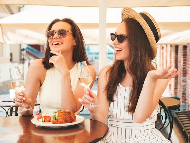 Deux jeunes belles femmes hipster souriantes dans des vêtements d'été à la modeFemmes insouciantes posant au café-véranda dans la rueModèles positifs buvant du vin blanc au chapeauProfitant de leurs vacances