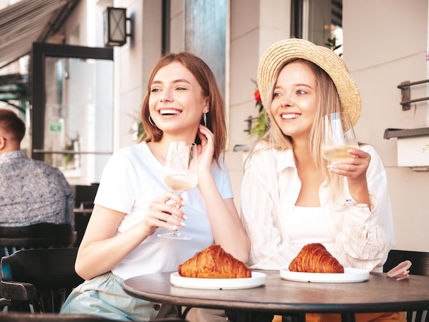 Deux jeunes belles femmes hipster souriantes dans des vêtements d'été à la modeFemmes insouciantes posant au café-véranda dans la rueModèles positifs buvant du vin blancProfitant de leurs vacancesManger un croissant