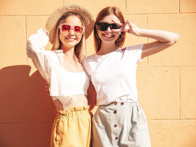 Deux jeunes belles femmes hipster souriantes dans des vêtements d'été à la modeFemmes insouciantes sexy posant près d'un mur blanc dans la rue Modèles purs positifs s'amusant au coucher du soleil étreignant et devenant fou