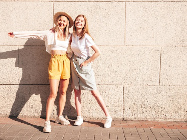 Deux jeunes belles femmes hipster souriantes dans des vêtements d'été à la modeFemmes insouciantes sexy posant près d'un mur blanc dans la rue Modèles purs positifs s'amusant au coucher du soleil étreignant et devenant fous