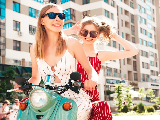 Deux jeunes belles femmes hipster souriantes en salopette à la modeFemmes insouciantes sexy conduisant une moto rétro sur le fond de la rueModèles positifs s'amusant à conduire un scooter italien classique en lunettes