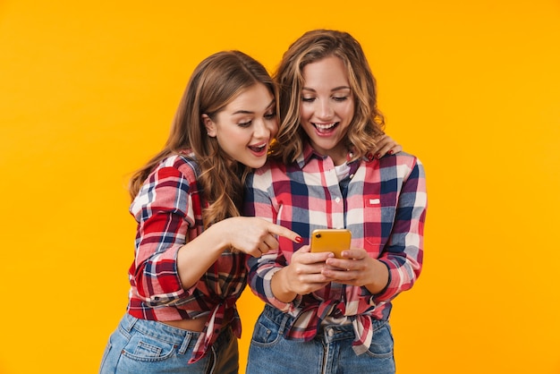 deux jeunes belles filles portant des chemises à carreaux souriant et utilisant un téléphone portable isolé
