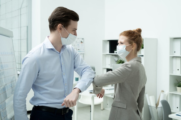 Deux jeunes chefs de bureau heureux en tenues de soirée et masques de protection se donnant un coup de coude au lieu de poignée de main ou de câlin