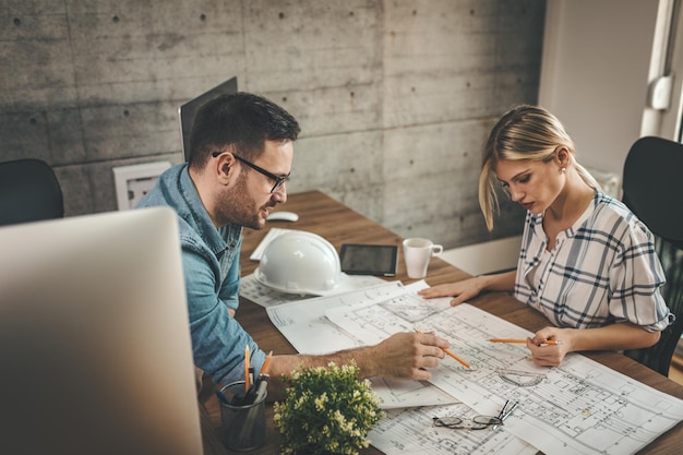 Deux jeunes collègues architectes pensifs travaillent sur un nouveau projet, analysent des plans et en parlent au bureau.