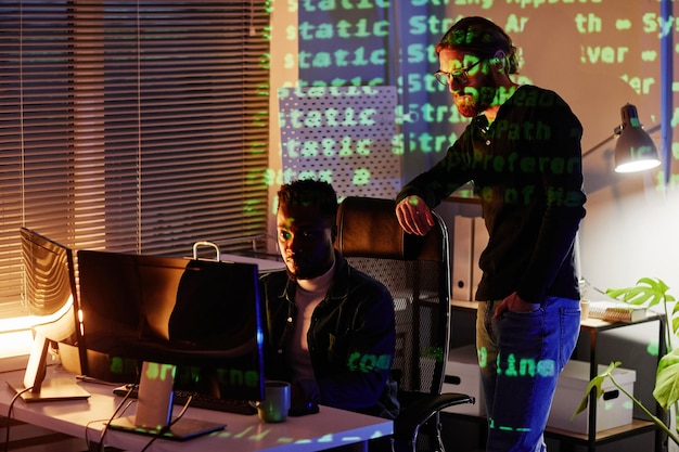 Deux jeunes développeurs de programmes interculturels regardant un écran d'ordinateur pendant que l'un d'eux est assis à son bureau pendant une réunion de nuit