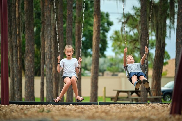 Deux jeunes enfants adolescents, une fille et un garçon, jouent ensemble sur des balançoires à l'extérieur lors d'une journée de vacances ensoleillée.