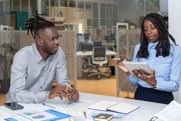 Deux jeunes entrepreneurs afro-américains avec une tablette numérique et des graphiques sur la table dans une salle de conférence tenant une réunion au bureau