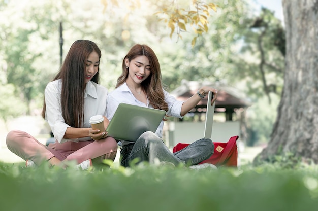 Deux jeunes étudiants universitaires asiatiques utilisant un ordinateur et parlant avec bonheur tout en étant assis sur l'herbe verte sous un grand arbre au parc.