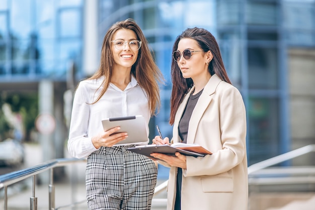 Deux jeunes femmes d'affaires avec tablette et ordinateur portable marchant à l'extérieur près d'un centre d'affaires moderne.