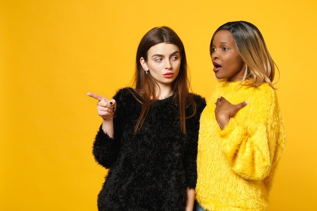 Deux jeunes femmes amies européennes et afro-américaines en vêtements jaunes noirs debout posant isolés sur fond de mur orange vif, portrait en studio. Concept de style de vie des gens. Maquette de l'espace de copie.