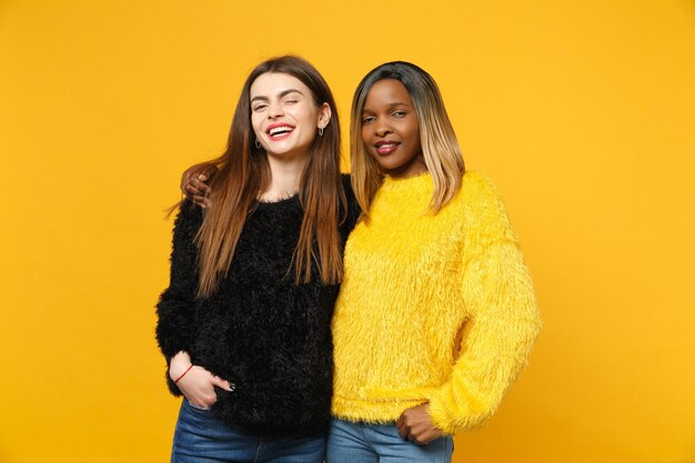 Deux jeunes femmes amies européennes et afro-américaines en vêtements jaunes noirs debout posant isolés sur fond de mur orange vif, portrait en studio. Concept de style de vie des gens. Maquette de l'espace de copie.