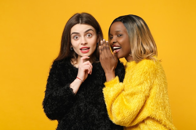 Deux jeunes femmes amies européennes et afro-américaines en vêtements jaunes noirs debout posant isolés sur fond de mur orange vif, portrait en studio. Concept de style de vie des gens. Maquette de l'espace de copie.