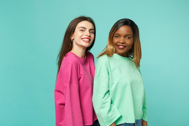 Deux jeunes femmes amis européens et afro-américains en vêtements verts roses debout posant isolés sur fond de mur bleu turquoise, portrait en studio. Concept de style de vie des gens. Maquette de l'espace de copie.