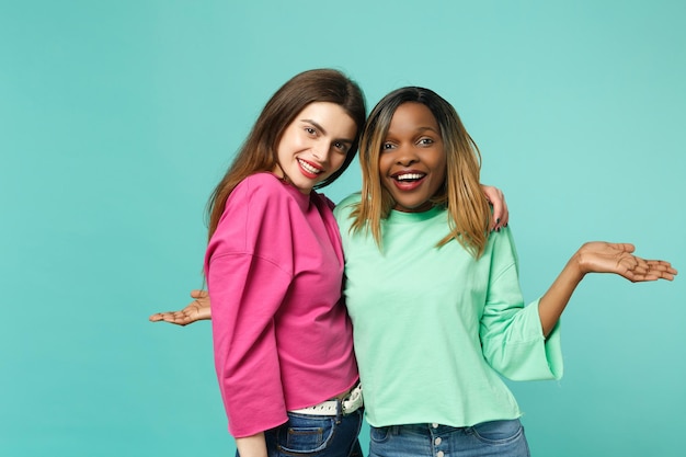 Deux jeunes femmes amis européens et afro-américains en vêtements verts roses debout posant isolés sur fond de mur bleu turquoise, portrait en studio. Concept de style de vie des gens. Maquette de l'espace de copie.