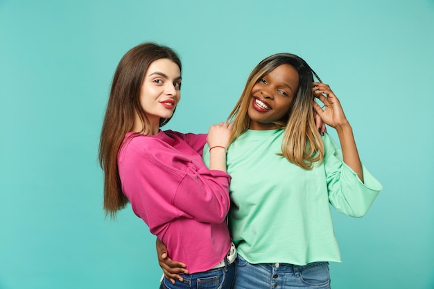 Deux jeunes femmes amis européens et afro-américains en vêtements verts roses debout posant isolés sur fond de mur bleu turquoise, portrait en studio. Concept de style de vie des gens. Maquette de l'espace de copie.