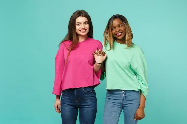 Deux jeunes femmes amis européens et afro-américains en vêtements verts roses debout posant isolés sur fond de mur bleu turquoise, portrait en studio. Concept de style de vie des gens. Maquette de l'espace de copie.