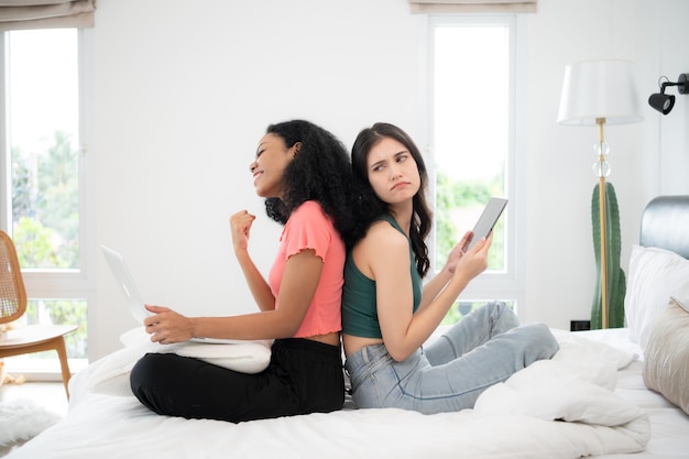 Deux jeunes femmes assises sur le lit dans la chambre et utilisant un ordinateur portable et une tablette pour jouer au jeu