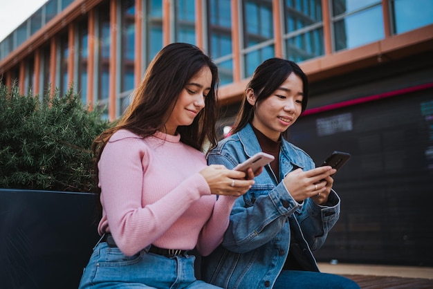 Deux jeunes femmes assises et utilisant leurs téléphones intelligents pour regarder leurs médias sociaux pendant qu'elles s'amusent