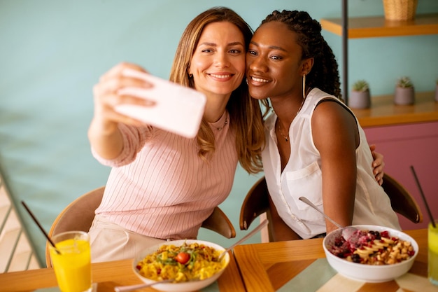 Deux jeunes femmes caucasiennes et noires prenant un selfie avec un téléphone portable dans le café