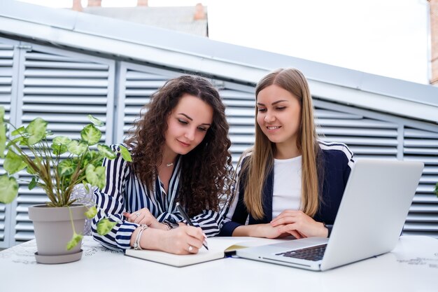 Deux jeunes femmes chefs d'entreprise prospères sont assises à une table avec un ordinateur portable et un ordinateur portable travaillant sur un nouveau projet de développement, des étudiantes rédigent un rapport sur le travail de leur ordinateur