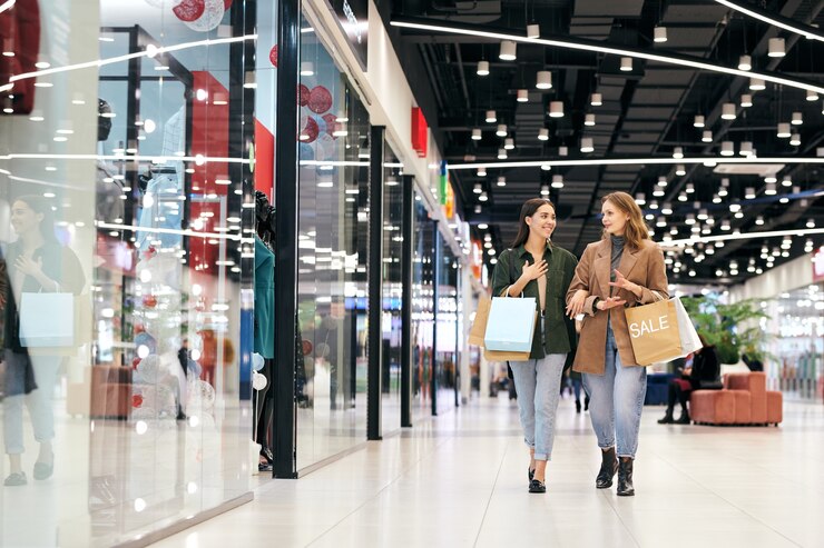 Photo deux jeunes femmes contemporaines en vêtements décontractés élégants marchant le long des vitrines à l'intérieur du centre commercial et discutant de ce qu'elles aimeraient acheter